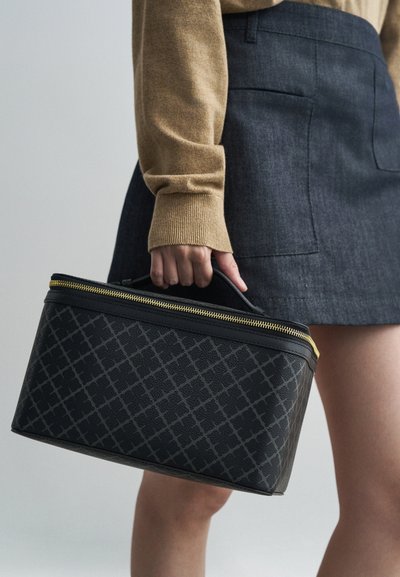 Melna teksturēta soma ar rāvējslēdzēja aizdari un zelta aksesuāriem, kas raksturo ģeometrisku rakstu. To tur persona, kas ģērbusies smilškrāsas džemperī un zilo džinsu svārkos.