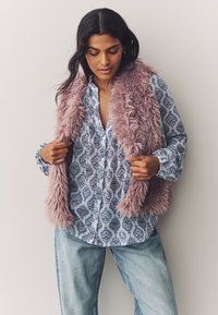 Faux fur vest in light pink over a blue patterned button-up shirt. The shirt features intricate designs, paired with relaxed denim jeans.