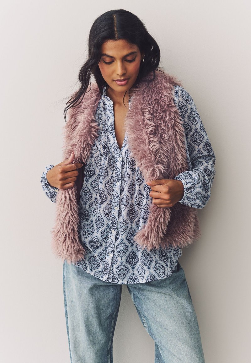 Faux fur vest in light pink over a blue patterned button-up shirt. The shirt features intricate designs, paired with relaxed denim jeans.