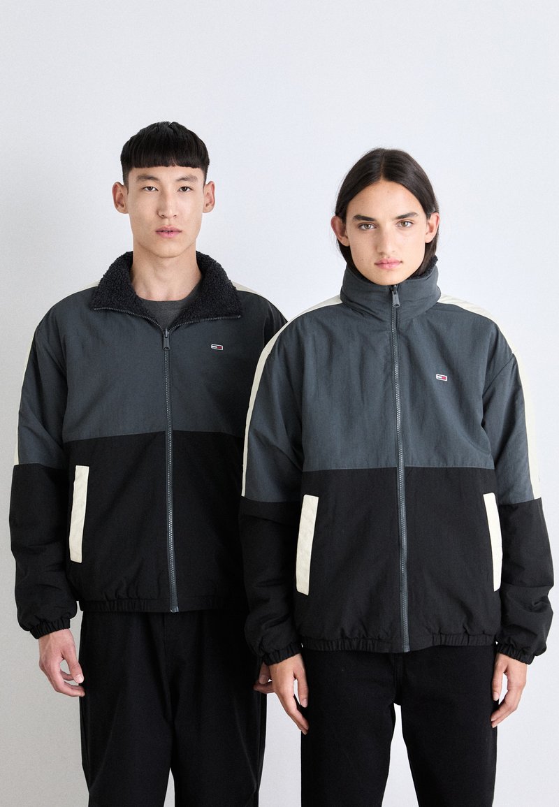 Dos personas llevan chaquetas con cremallera en tonos de gris y negro, con un cuello texturizado, puños elásticos y paneles laterales en contraste.