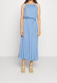 Helles blaues, ärmelloses Maxikleid mit gerafftem Ausschnitt und Taillengebund, kombiniert mit grünen Riemchensandalen. Glatter Stoff, luftiges Design.