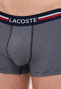 Gestreifte Herrenboxershorts in Marineblau und Weiß mit einem einfarbigen Marineblauen Bund, auf dem das "LACOSTE"-Logo in Weiß steht. Glattes Material mit genähten Nähten.