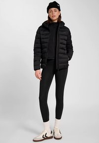 Zwarte donsjas met capuchon, ritssluiting en doorgestikt ontwerp. Draagt over een zwarte top en legging, gecombineerd met crèmekleurige en bruine sneakers.