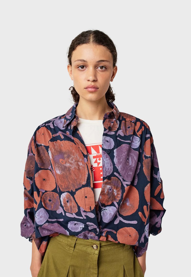 Giovane donna con capelli ricci indossa una camicia oversize blu navy con grandi stampe floreali arancioni e viola sopra una maglietta bianca con grafica e pantaloni verdi.