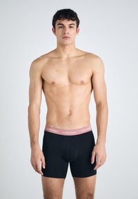 Fekete pamut boxer alsónadrág rózsaszín Calvin Klein derékkal, kényelmes fazonnal és sima textúrával. A modellen félmeztelen látható.