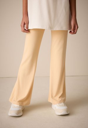 Person iført beige udvidede bukser, hvide sneakers og en cremefarvet lang top, stående på en lys gulv mod en neutral baggrund.