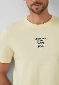 Gelbes Baumwoll-T-Shirt mit Rundhalsausschnitt, das schwarzen gestickten Text trägt: "LONESOME RIDER SOCIAL Club." Einfaches Design mit kurzen Ärmeln.