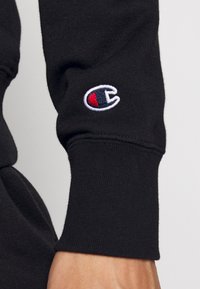 Polsino di una felpa nera con un logo ricamato multicolore con accenti rossi, bianchi e blu. Il tessuto sembra morbido e testurizzato.