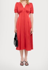 Mujer vistiendo un vestido midi rojo con botones forrados de tela, mangas abullonadas, sosteniendo un bolso negro y usando tacones negros de tiras.