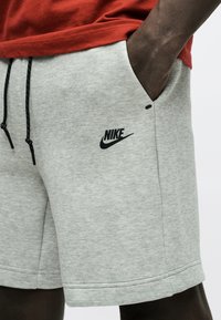 Κοντινό πλάνο ατόμου που φορά γκρι ανοιχτό σορτς Nike με μαύρα κορδόνια και το χέρι στην τσέπη, συνδυασμένο με κόκκινο πουκάμισο.