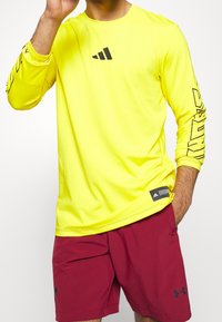 Camiseta atlética de manga larga, de un brillante color amarillo, con branding negro en el pecho y la manga; combinada con pantalones cortos color burdeos. Tela suave y elástica.