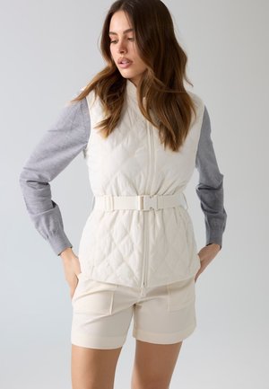 Kobieta nosząca biały pikowany kamizelkę z paskiem na szarej bluzce z długim rękawem i kremowych szortach, stoi z rękami w kieszeniach.