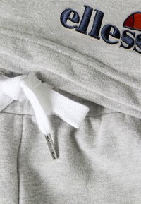 Pantalon de survêtement gris clair avec cordon blanc noué en un nœud et une ceinture élastique partiellement visible brodée de "ellesse" en bleu marine et orange.