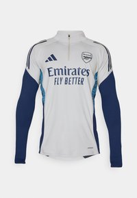 Hvid langærmet trøje med marineblå detaljer, halv-zip halsudskæring, med "Emirates Fly Better" logo og Arsenal klublogo.