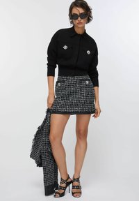 Giacca corta nera a maglia con due tasche frontali e gemme decorative; abbinata a una mini gonna in tweed bianco e nero e tacchi neri con cinturini.