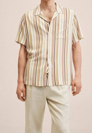 Mężczyzna noszący koszulę na krótki rękaw zapinaną na guziki z pionowymi żółtymi, brązowymi i beżowymi paskami, zestawioną z jasnobeżowymi spodniami.