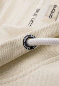 Gros plan sur un sweat à capuche beige montrant un cordon blanc passant à travers un œillet métallique noir marqué "ombre wear" et une partie de texte imprimé.