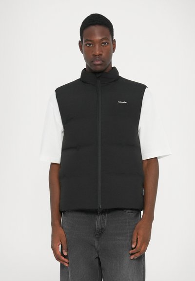 Gilet puffo nero con colletto alto, chiusura con zip e logo sul lato sinistro del petto. Indossato sopra una camicia bianca a maniche corte. Sfondo neutro.