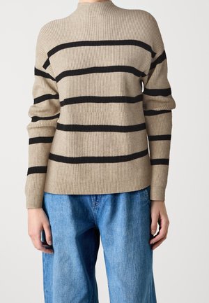 Beige ribstrikket sweater med sorte horisontale striber båret med løse blå denimbukser, vist fra nakke til hofter.