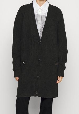 Personne portant un long cardigan noir boutonné sur une chemise blanche à col avec un panneau plissé gris, associé à un pantalon noir.