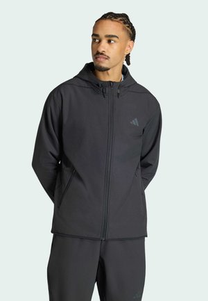 Mann mit geflochtenem Haar, der eine schwarze Kapuzenjacke mit Reißverschluss und passende Hose trägt, schaut nach links vor hellem Hintergrund.