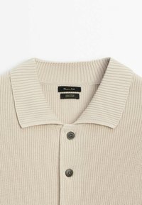 Chemise en coton beige tricotée avec col large côtelé et deux boutons foncés sur le devant.