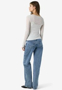 Sieviete, kas uzvilkusi gaiši pelēku garām piedurknēm topu ar spageti siksnām, kas kārtojams virsū, zilos platās kājas džinsos un melnās kurpēs, skatās sānis.