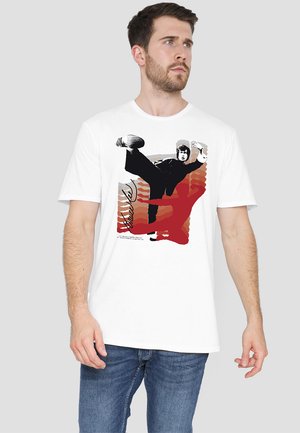 Man wearing white T-shirt with graphic of a person performing a high kick over a red and brown abstract background.