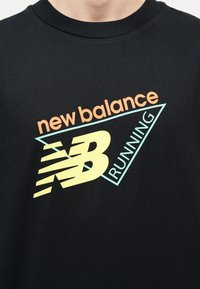 Κοντινό πλάνο μαύρου πουκάμισου με πολύχρωμο λογότυπο New Balance Running που παρουσιάζει ένα στυλιζαρισμένο "NB" και σχεδίαση τριγώνου.