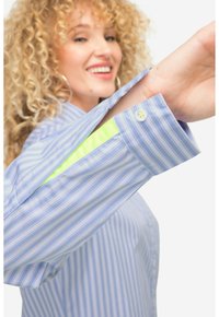Camisa de rayas azul claro con mangas largas, puños de botón y un acento verde neón en el interior de la manga. La tela es suave y ligera.