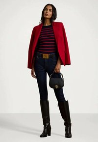 Γυναίκα φοράει κόκκινο σακάκι πάνω από ριγέ μπλούζα, σκούρα τζιν και μαύρες μπότες μέχρι το γόνατο, κρατώντας μία μικρή μαύρη τσάντα με χρυσό κούμπωμα.