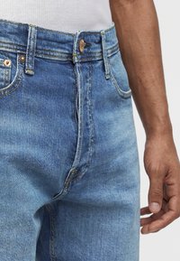 Κοντινό πλάνο από τζιν μεσαίου μπλε χρώματος με μπροστινό κουμπί και φερμουάρ, φορεμένα από ένα άτομο με λευκό πουκάμισο, που δείχνει το δεξί χέρι χαλαρό δίπλα στο σώμα.