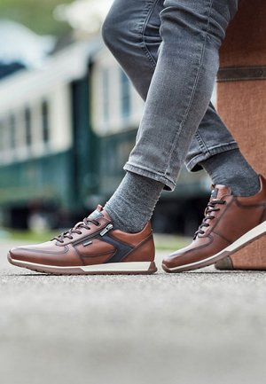 Personne portant un jean gris et des chaussettes grises, assise les jambes croisées, portant des baskets en cuir marron décontractées avec des fermetures éclair latérales.