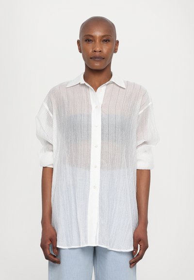 Cămașă albă cu mânecă scurtă, tip buton, cu un design texturat și transparent. Are un guler clasic și nasturi vizibili, asortată cu pantaloni din denim deschis.
