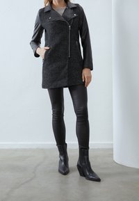 Černý kabát z vlněné směsi s koženými rukávy, asymetrickým zapínáním na zip, dvěma bočními kapsami a černými kotníkovými botami na vysokém podpatku.