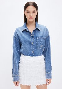 Camicia di denim con taschino frontale e dettagli scuciti, abbinata a una mini gonna bianca texture con dettagli sfrangiati e tessuti sovrapposti.