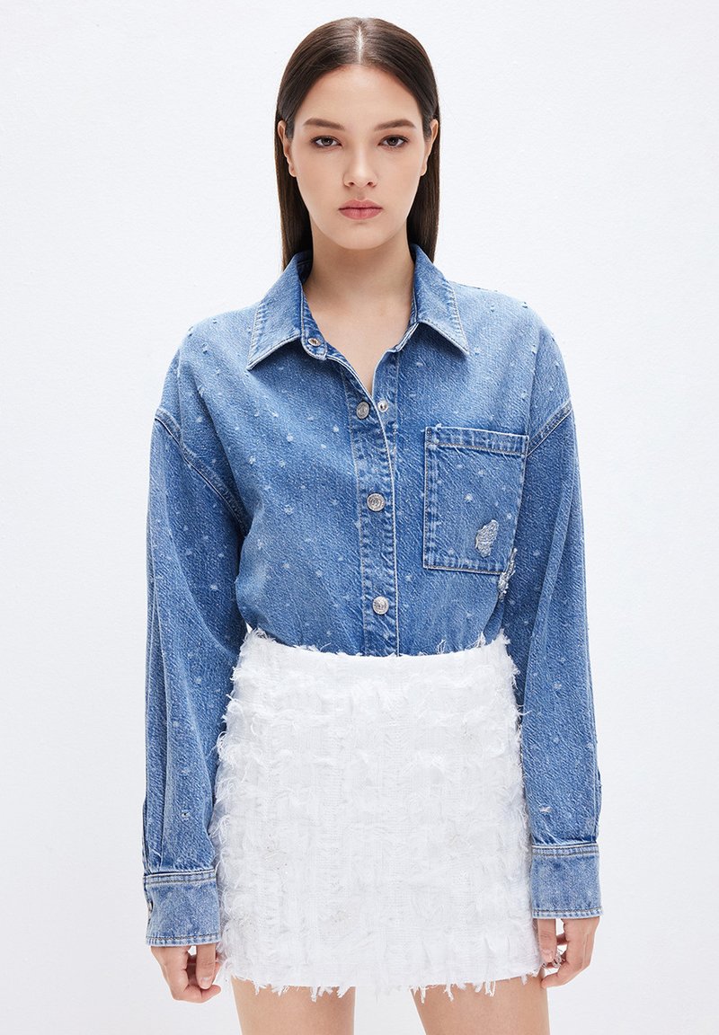 Camicia di denim con taschino frontale e dettagli scuciti, abbinata a una mini gonna bianca texture con dettagli sfrangiati e tessuti sovrapposti.