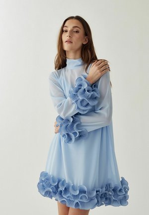 Kvinde iført en gennemsigtig lyseblå kjole med høj hals og 3D-rufflede detaljer på ærmer og skørt, stående foran en ensfarvet hvid baggrund.