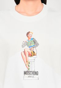 T-shirt blanc avec un design graphique coloré représentant une femme assise en haut rayé, accompagné du texte "MOSCHINO JEANS" en dessous.