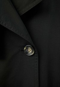 Gros plan sur le col d'une veste noire avec surpiqûres visibles et un seul bouton style écaille de tortue fermé.