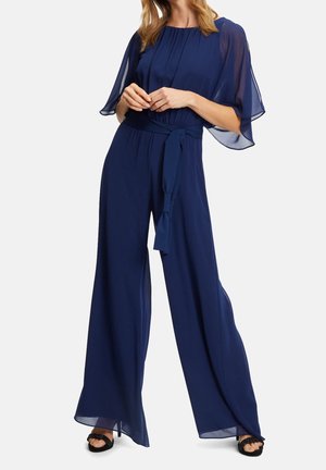 Femme portant une combinaison bleu marine à jambes larges avec des manches fluides transparentes et une ceinture à la taille, associée à des talons ouverts noirs.