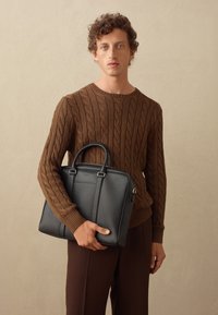 Maletín de cuero negro con dos asas, que presenta costuras sutiles y una textura suave, sostenido por una persona que lleva un suéter de punto trenzado marrón.