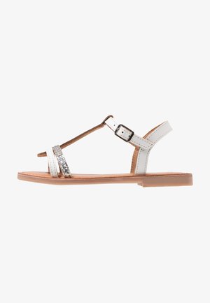 Sandalias planas blancas con diseño de tira en T, tira plateada adornada y cierre de hebilla. Parte superior de piel, plantilla texturizada y suela marrón claro.