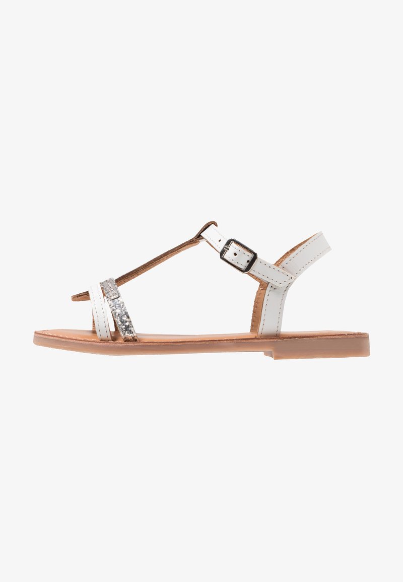 Sandalias planas blancas con diseño de tira en T, tira plateada adornada y cierre de hebilla. Parte superior de piel, plantilla texturizada y suela marrón claro.