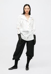 White oversized shirt with textured detailing, buttons, and black low-rise trousers. Model wearing black gloves and fitted socks.