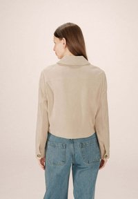 Donna con lunghi capelli castani, indossa una giacca cropped beige e jeans a vita alta blu, in piedi di spalle contro uno sfondo semplice.