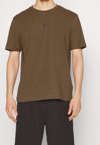 Brun bomull t-shirt med korta ärmar, rund halsringning och diskret logotyp. Bärs med mörka shorts som har en avslappnad passform och mjuk textur.