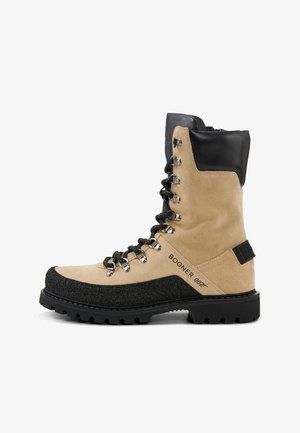 Wysokie do kolan buty trekkingowe wykonane z beżowego zamszu z czarnymi skórzanymi akcentami. Posiadają metalowe dziurki, teksturowaną czarną gumową podeszwę oraz detale z logo.