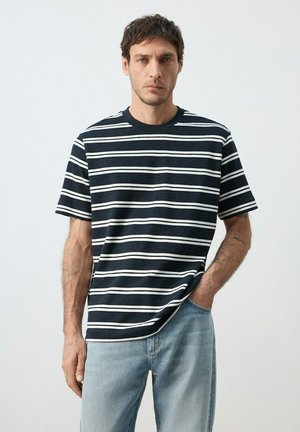 Hombre con cabello corto y oscuro, vestido con una camiseta de rayas blancas y negras y jeans azul claro, de pie frente a un fondo liso.