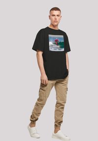 Camiseta negra de algodón con un gráfico cuadrado de un surfista en vibrantes tonos de rosa y verde, combinada con pantalones cargo beige y zapatillas blancas.